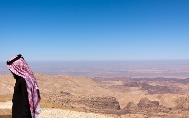 A young sheikh (with his traditional red/white Keffiyeh) in front of a desert landscape which may symbolize a field of fossil oil or the oil industry.