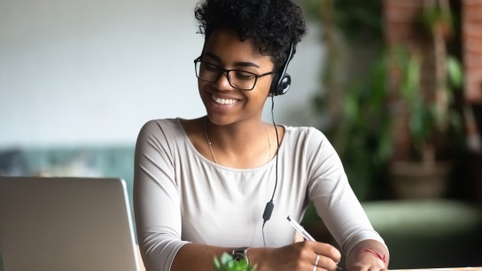 Smiling african American millennial female student in headphones and glasses sit at desk watch webinar making notes, happy biracial young woman in earphones work study using computer write in notebook