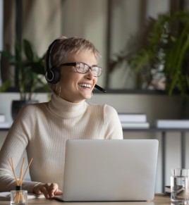Overjoyed young caucasian businesswoman in headphones glasses have fun talking on video call on laptop at workplace. Smiling female employee in earphones laugh enjoy watching webinar on computer.