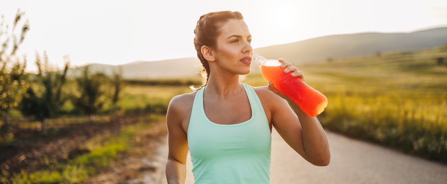 Attractive young blonde sportswoman is refreshing with cold drink after jogging in nature on sunny summer day at sunset
