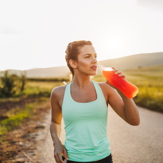 Attractive young blonde sportswoman is refreshing with cold drink after jogging in nature on sunny summer day at sunset