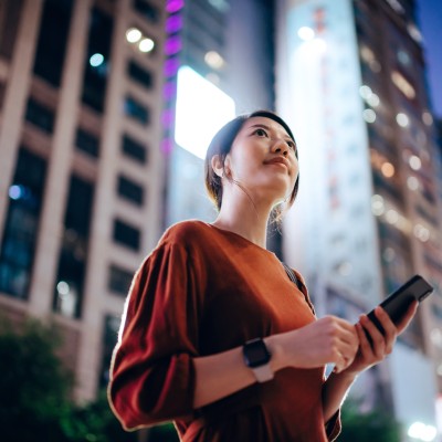 Low angle portrait of confident and beautiful young Asian businesswoman using smartphone while commuting in downtown city street, looking up to the sky against illuminated urban skyscrapers in the city.