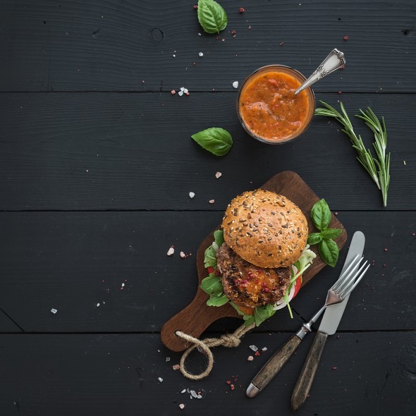 Fresh homemade burger on dark serving board with spicy tomato sauce, sea salt and herbs over dark wooden background. Top view, copy space
