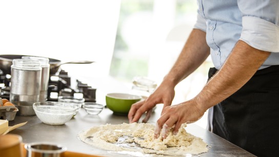 Mid adult man with black apron baking pizza dough in the kitchen.