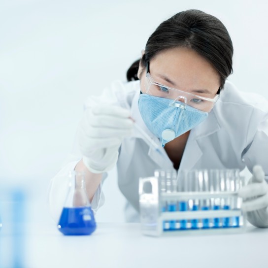 Asian woman scientist holding several test tubes and filling them with blue chemical by dropper, she is wearing safety goggle, glove, protective mask and lab coat.