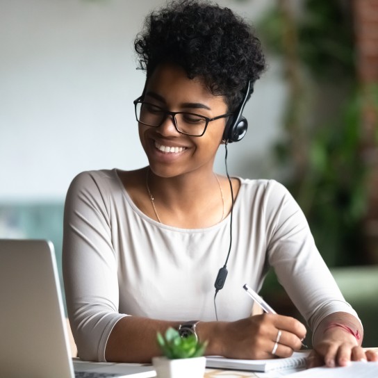 Smiling african American millennial female student in headphones and glasses sit at desk watch webinar making notes, happy biracial young woman in earphones work study using computer write in notebook