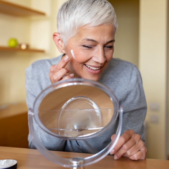 Beautiful senior woman checking her face skin and looking for blemishes. Portrait of mature woman massaging her face while checking wrinkled eyes in the mirror. Wrinkled lady with grey hair checking wrinkles around eyes, aging process.