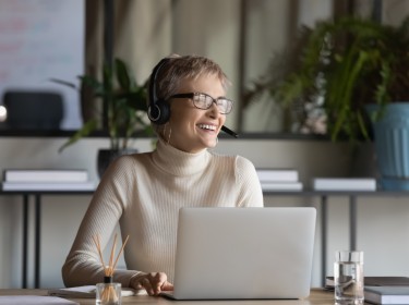 Overjoyed young caucasian businesswoman in headphones glasses have fun talking on video call on laptop at workplace. Smiling female employee in earphones laugh enjoy watching webinar on computer.