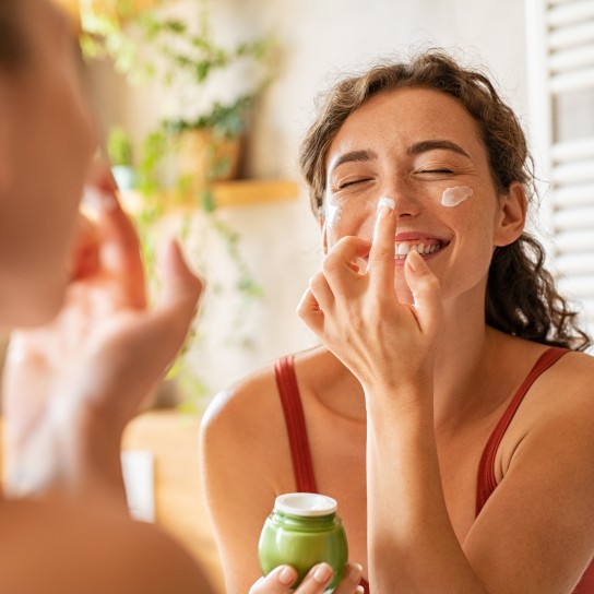Playful young woman applying cream on nose. Cheerful girl holding green lotion jar standing in front of mirror applying moisturizer on nose. Beautiful woman taking care of skin by applying moisturizer every day in the morning.