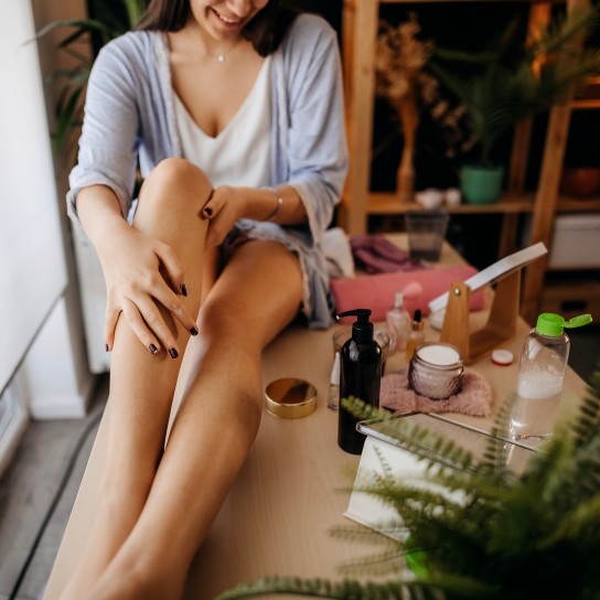 Woman sits on a table in her bedroom and nourishes her feet with cream