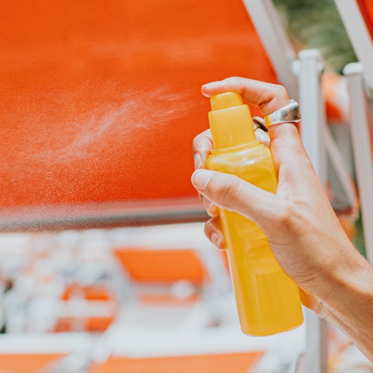 Close-up of a woman's hand pressing down on a nozzle of a liquid sunscreen container, spritzing it onto her friend's back.