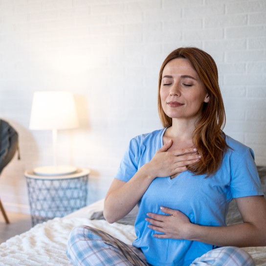 Young woman sitting in bed with eyes closed, hands on chest and abdomen, practicing mindful breathing for relaxation and stress relief in her cozy bedroom
GettyImages-2207000026.jpg