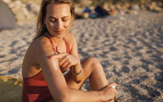 A smiling young woman wearing a one-piece swimsuit sitting on the beach and applying sunscreen on a sunny summer day at the seaside
GettyImages-2169631007.jpg